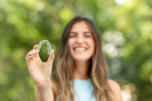 Smiling woman holding clear Invisalign aligner outdoors, showcasing orthodontic treatment options for underbite correction.